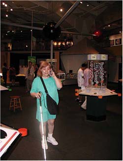 Image shows a woman navigating on the exhibit floor a the New York Hall of Science. She carries a long cane in one hand and a cell phone in the other. 