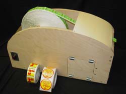 Image shows a wooden prototype with a large diameter roll of stickers. The sides of the prototype are rounded on the top to reduce sharp outward corners. The hinged access door and power switch are shown on the right side product. A small optical sensor is located under the area where the sticker is exported. Also shown in this image is a selection of the different sticker sizes and geometries. 