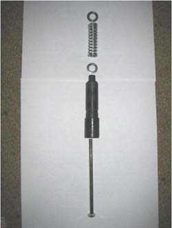 Image shows the five components that make up the shock absorber system. There is a spring, with a washer on each side. Next is a plastic molding piece. The top of the molding fits inside the bottom of the spring, acting as the bottom surface on which the spring compresses. Finally, there is a long screw with a hexagonal head. The hex screw fits up inside of the plastic molding and through the spring (and both washers).  