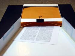Image shows the frontal view of the preliminary design prototype that is constructed out of cardboard. The funnel shaped aligner tray leads into an adjoining envelope holder. The envelope holder has an overhead ledge mounted over one end to which the non-flapped side of a 10 x 13 envelope is secured with a binder clip. This provides an elliptical envelope opening. At the other end of the holder, there is a restraining ledge to keep the envelope in place. The envelope rests on a tray, which is biased by a resilient spring underneath (not shown in photo). A document is resting on the tray surface and is half-inserted into the envelope.