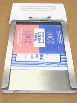Image shows the top view of the 9.5 x 12.75 envelope inserting aid with mail material loaded on the aligner tray and an envelope secured over the channel by a clip. The 16 x 11.3 aluminum aligner tray has two perpendicular lipped edges and mail material fits under the lip. The top ledge of the channel slopes down, thus the envelope slants downwards from the channel towards the table surface. The channel defines an 11.3 x 0.7 rectangular document feed path from the aligner tray into the open envelope. 