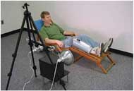 This image shows a researcher reclined in a lounge chair with the erythema inducer strapped to his lower right leg. A camera and two lights are aimed at the erythema site.