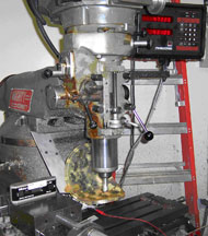 This illustration shows the HPM positioned in the device-grip of a manual mill tilted to the desired angles to establish a vertically orthogonal position of the probe with the associated test point. Adjacent to the HPM shell is a digital protractor used to precisely measure angles to appropriately access test points. There are voltage supply and serial communication lines connected to the HPM to power the device and obtain the calculated output data. 