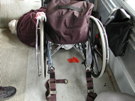 Post-crash photo taken from behind the wheelchair in case WC-006 showing the deceased occupant lying on the floor of the van in front of the wheelchair.  The deceased occupant’s head is toward the center of the van and the occupant is lying on her left side.