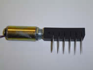 Image shows the side view of the solenoid attached to a pick rod containing commercial picks. The tubular solenoid is 2” long with a 1” diameter. A metal plunger runs through the middle of the solenoid and connects to the pick rod. The Delrin plastic pick rod is a rectangular block with six slits, into which six commercial picks have been inserted. 
