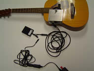 Image shows the overhead view of the entire device with connections. An AC/DC converter is just visible and is connected to a rectangular pedal through a 1/8” mono connection. The pedal is then connected to the strumming component through ¼” stereo connections. The strumming component is mounted to the guitar face.