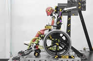 This side view photo shows a Hybrid three, six year old ATD seated in a manual pediatric wheelchair. The wheelchair is equipped with a headrest. The wheelchair is secured to a test sled with four strap-type tiedowns. The ATD is wearing a three-point occupant restraint. There is whit arrow on the photo pointing to the joint where the anterior-posterior headrest stem meets the vertical headrest stem.