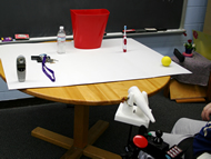 This photo shows the set of objects: a cordless phone, keys, water bottle, trash bin, toothbrush, and ball (from left to right in the picture). The objects were arranged on a horizontal surface. The participant was positioned in front of the table, approximately centered. The laser joystick was mounted to his/her wheelchair. 