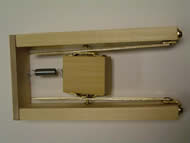 Image shows the overhead view of the mechanical track demonstration of principle constructed using a wooden skeleton with metal components. Two ½” X 2” X 10” pieces of poplar wood serve as the track support pieces with a sliding track attached to each one. A smaller wooden piece, the pick-holding component, lies in between the tracks suspended through screw connections. A spring links the pick-holding component to a wooden end piece, perpendicularly attached to both track support pieces. 