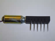 Image shows the side view of the solenoid attached to a pick rod containing commercial picks. The tubular solenoid is 2” long with a 1” diameter. A metal plunger runs through the middle of the solenoid and connects to the pick rod. The Delrin plastic pick rod is a rectangular block with six slits, into which six commercial picks have been inserted.  