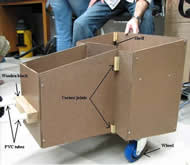 Image shows the side view of the preliminary spatial prototype that is constructed out of tempered hardboard.  Two PVC pipe attachment tubes are inside a wooden block that is attached to the middle of front of the cart.  The device, gradually increasing in height as the distance increases from the attachment point, consists of a thinner and longer front chamber followed by a wider and shorter rear chamber in which a shelf is housed.  Small wooden blocks serve as corner joints by which nails and screws secure the hardboard attachments.  Two swivel, rubber-coated caster wheels are beneath the rear chamber close to the sides of device. 