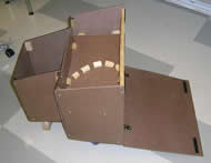 Image shows the top view of the functional prototype that is constructed out of tempered hardboard.  The device’s shape mirrors the spatial prototype with a few adjustments.  The front chamber is shortened significantly and the shelf is lowered in the rear chamber.  A wooden beam with Velcro® pieces towards the ends attaches the rear side panels near the top.  The rear hardboard piece is an adjustable flap that is on the ground and connected to the main chamber by two brass hinges.  Two Velcro® pieces align the sides of the flap such that it can connect to a wooden cross beam.  A series of small wooden blocks on the shelf create supports around where a watering pail could be placed. 