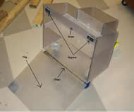 Image shows a rear view of the nearly complete device composed of Lexan®, a GE-brand polycarbonate, with its flap in the down position.  Nearly identical to the functional prototype, the device is characterized by a frosty shower door-like finish.  Magnets protrude from the sides of the beams, attracting metal plates on the flap.  A hinge extends the length of the bottom and swivel wheels are beneath the main chamber.  The 0.375” Lexan® pieces are attached via stainless steel screws that are directly screwed into the thickness of the sides.  The Lexan® sides are transparent enough to see the lengths of these screws even though they cannot be touched anywhere besides on their heads.  A 0.22” thick polycarbonate shelf sits atop Lexan® support blocks. 