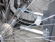 Image shows the attachment mechanism close-up.  A Lexan® block consisting of three 0.375” Lexan® slabs connected with a silicone-based adhesive comprises the side attached to the device.  Two aluminum rods 0.625” in diameter are inlayed inside the block near the ends and are fitted inside tubular openings in the wheelchair.  Stainless steel screws from the top and bottom of the block go through to the tubes to secure their attachments.  In the center, a hinge is attached to the top of the Lexan® block as well as the back of a small white plastic block.  A rubber-coated metal hook is screwed into the plastic block, which then grabs onto the wheelchair’s axle. 