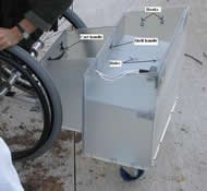 Image shows a side view of the final device composed of Lexan®.  The device is the exact same in structure to Photo 5 with some additional features.  A handle near the front of the device allows users to easily attach the device to a wheelchair.  The attachment mechanism is connected to the front of the device and the shelf has an increased thickness of 0.375” as well as an ergonomic handle.   Two hooks on the side of the device can be used to store the shelf, which has two holes to place over the rounded metal hooks.   