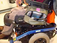 This photograph shows the prototype attached to our client’s wheelchair with the zippered bag open to reveal its contents. The zippered bag attached to our client’s left armrest contains the plastic container housing the pump, battery, and semi-clear white extension tubing. The black extension tubing coming out of this bag has been routed along his wheelchair, attached via cable clamps, and connected to the drainage tube of his leg bag located medially under his pants near his left ankle.