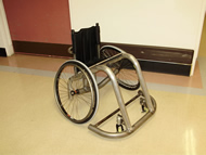 This photograph shows the wheelchair in an isometric view.  It shows two side frames weld to the front bumper, and that the axle connects to the side frames by two vertical pipes. 
