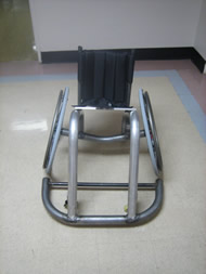 This photograph shows the wheelchair in a front view.  It shows how the bumper connects to the side frame, which provides a mount for the front casters and the 15 degree outward camber of each drive wheel. 