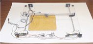 The picture shows vertically oriented motorized lead screws on either side of a wooden board, a third motorized lead screw connects between the two horizontal lead screws. A small piece of conductive foam is attached to the screw nut which moves along the horizontal axis. 